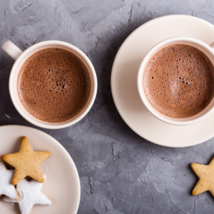 two mugs of hot chocolate, one on a saucer, next to a plate of three star cookies with one star cookie on the table